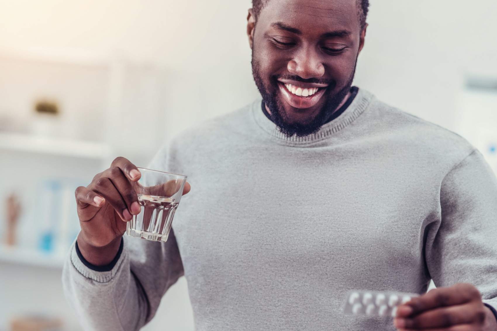 Such a relief. Beaming millennial guy in casual looking a blister with pills while holding a glass of water and taking his prescription.