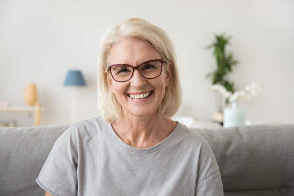 Smiling middle aged mature grey haired woman looking at camera, happy old lady in glasses posing at home indoor, positive single senior retired female sitting on sofa in living room headshot portrait